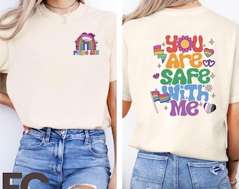 May include: A white t-shirt with a rainbow and heart design that says "Pride Ally" on the front and "You are safe with me" on the back. The shirt is shown on a model wearing blue jeans.
