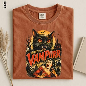 May include: A rust-colored short-sleeve t-shirt featuring a graphic print of a black cat with glowing orange eyes, a haunted house, bats, and a woman in a red dress. The text reads "NIGHT OF THE VAMPURR" in a bold, distressed orange font.