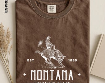 May include: An espresso-colored t-shirt featuring a white graphic of a person riding a bucking horse. The text reads EST 1889, MONTANA, and TREASURE STATE. The shirt has a Comfort Colors label at the inner neckline and a ribbed crew neck collar.