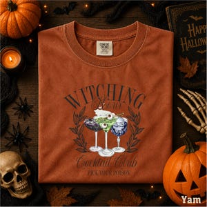 a t-shirt with a Halloween-themed graphic, including a witch's hat, cocktail glasses, and a skull, surrounded by Halloween decorations such as a pumpkin, bats, and a candle.