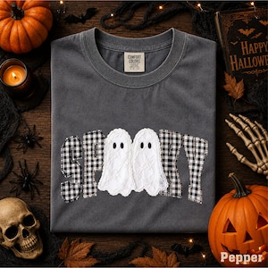 May include: A charcoal gray t-shirt featuring the word SPOOKY in a black and white buffalo plaid pattern, with two white lace ghosts replacing the letter O. The shirt has a crew neckline and a Comfort Colors brand label on the inside collar.