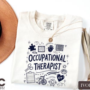 May include: Ivory-colored t-shirt with the words "Occupational Therapist" in navy blue, surrounded by related doodles. The shirt has a crew neck and short sleeves. A gold necklace with a pendant is draped on the shirt.