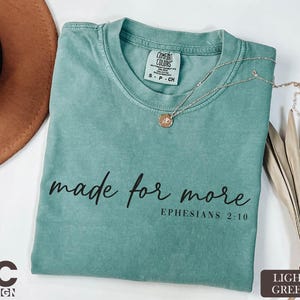 A turquoise t-shirt with the text "made for more" printed on it, along with a necklace and a brown hat, all placed on a white surface.