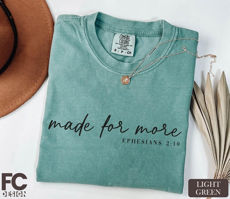 A turquoise t-shirt with the text "made for more" printed on it, along with a necklace and a brown hat, all placed on a white surface.