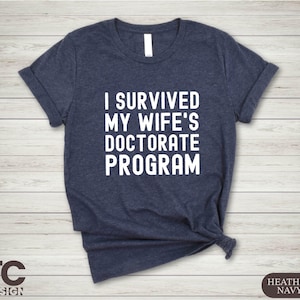 May include: A navy blue t-shirt with white text that reads "I survived my wife's doctorate program".