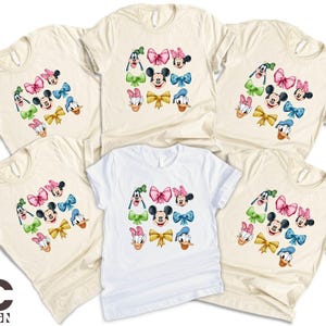 May include: Set of five cream-colored t-shirts with a colorful cartoon design of Mickey Mouse and Minnie Mouse characters. Each shirt features a different combination of the characters with their signature ears, gloves, and shoes.