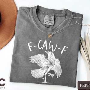 Gray t-shirt with white text and drawing of a bird, along with a necklace and a hat, all placed on a white surface.