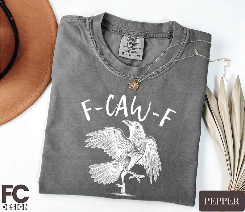 Gray t-shirt with white text and drawing of a bird, along with a necklace and a hat, all placed on a white surface.