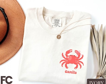 May include: Ivory-colored t-shirt with a red crab graphic and the name "Camila" in a matching font. The shirt has a crew neck and short sleeves. A gold necklace with a pendant is draped across the shirt.