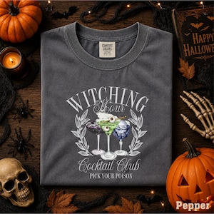a gray t-shirt with a Halloween-themed design, including a cocktail glass with a skull and crossbones, surrounded by autumn leaves and a pumpkin.