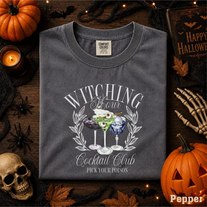 a gray t-shirt with a Halloween-themed design, including a cocktail glass with a skull and crossbones, surrounded by autumn leaves and a pumpkin.