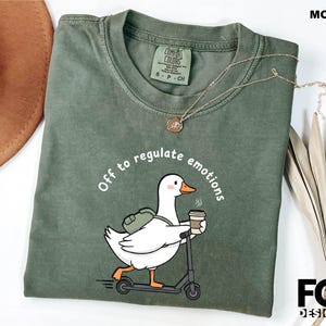 May include: Moss green t-shirt with the text "Off to regulate emotions" and a cartoon duck riding a scooter while holding a coffee cup. The shirt has a crew neck and short sleeves. A gold necklace is draped across the shirt.