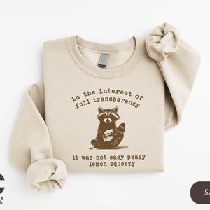 May include: Sand-colored sweatshirt with a raccoon graphic and the text "in the interest of full transparency, it was not easy peasy lemon squeezy." The sweatshirt has long sleeves and a crew neck.