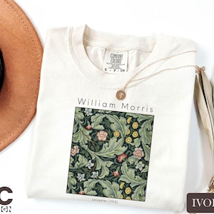 May include: Ivory-colored t-shirt featuring a William Morris floral design. The design includes green leaves, pink and yellow flowers, and the text "William Morris" and "Leicester (1912)". A gold necklace and a brown hat are also visible.
