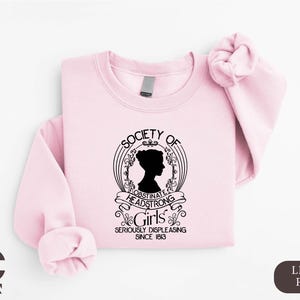 May include: Light pink sweatshirt with the text "Society of Obstinate Headstrong Girls" and a silhouette of a woman's profile. The text "Seriously Displeasing Since 1813" is also included. The sweatshirt has long sleeves.