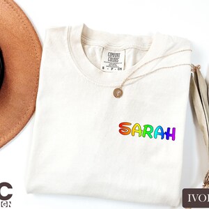 May include: Ivory-colored t-shirt with the name "Sarah" in rainbow-colored letters. The shirt has a crew neck and short sleeves. A gold necklace with a pendant is draped across the shirt. The shirt is made by Comfort Colors.