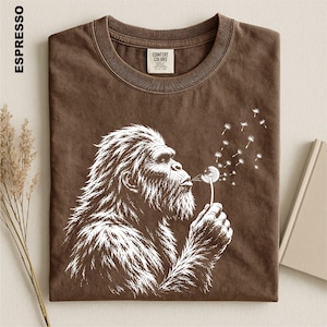a brown t-shirt with a white illustration of a gorilla blowing a dandelion, along with some dried flowers and a book.