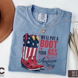 May include: A light blue t-shirt with a graphic of red cowboy boots with a US flag design. The text reads "We'll put a boot in your ass, it's the American way." A gold necklace and a brown hat are also visible.