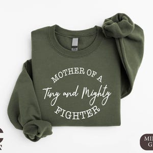 May include: A green sweatshirt with the text "Mother of a Tiny and Mighty Fighter" in white.