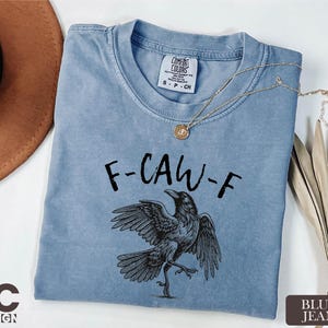 A blue t-shirt with a graphic of a bird and the text "F-CAW-F" printed on it, along with a necklace and a hat.