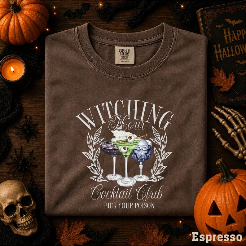 a t-shirt with a graphic design of cocktail glasses and a witch's hat, surrounded by Halloween decorations such as a pumpkin, bats, and a skeleton.