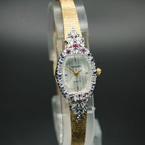 May include: A gold-toned Gruen quartz watch with a rectangular face, surrounded by a bezel of small, clear and red gemstones. The watch has a gold-toned band. The watch face is white with gold hands and markers.