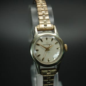 May include: A vintage Eterna-Matic wristwatch with a gold-tone band and a silver-tone case. The watch face is white with gold-tone hands and markers. The watch is displayed against a dark background.