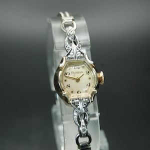 May include: A silver-toned Bulova wristwatch with a cream-coloured dial featuring black Arabic numerals. The watch case and the integrated bracelet links are accented with small clear stones. A gold-toned winding crown is on the right side.