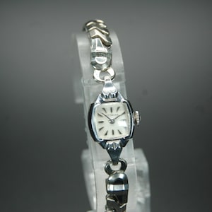 May include: A silver wristwatch with a square face and a silver metal band. The watch face has black Roman numeral hour markers and a silver second hand.