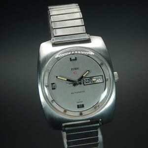 May include: A vintage Elgin automatic wristwatch with a silver-tone case and band. The watch face is light gray with black hands and markers, and a day-date display. The watch is set to Monday, the 20th.