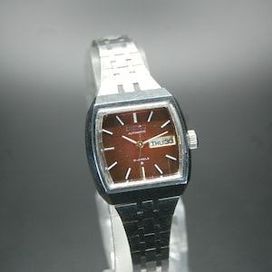 Könnte beinhalten: Vintage Automatik-Uhr mit rechteckigem braunem Zifferblatt und silberfarbenem Metallarmband. Das Zifferblatt zeigt Tag und Datum an. Die Uhr hat eine silberne Krone und ein Armband im Glieder-Stil.