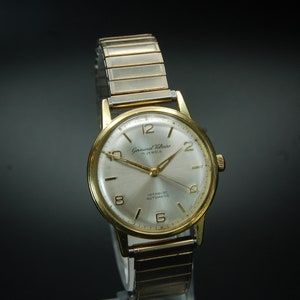 May include: A gold-tone wristwatch with a white face and black hands. The watch has a gold-tone band and says "Gerald Williams 17 Jewels Incabloc Automatic" on the face.