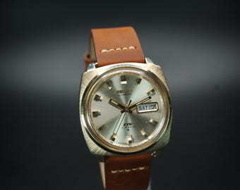 May include: A vintage Seiko automatic watch with a gold-toned case and a brown leather strap. The watch face is silver with gold hands and markers, and the day is displayed as "SAT 7".