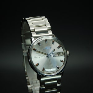 Puede incluir: Un reloj de pulsera plateado con una esfera blanca y una correa de metal plateado. La esfera del reloj tiene una ventana de fecha que dice "SÁB 2" y las palabras "Ocean Star 20+2804" impresas en ella. La marca del reloj es "Alpina".