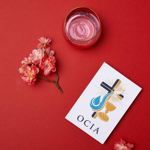 Puede incluir: Imagen de fondo rojo con un vaso de l&iacute;quido rosa, una ramita de flores rosas y una tarjeta blanca con un gr&aacute;fico religioso y la palabra OCIA. El gr&aacute;fico de la tarjeta incluye una cruz, una paloma, un c&aacute;liz y pan.