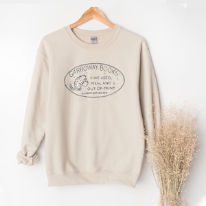 Può includere: Felpa beige con grafica ovale nera con la scritta "GARROWAY BOOKS" e il testo "FINE USED, NEW, AND OUT-OF-PRINT - CLOSED SATURDAYS". La felpa è appesa a una gruccia di legno.