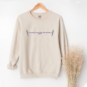 Może przedstawiać: Beżowa bluza z fioletowym napisem "I'm going to ask again. Who did this to you?" i ilustracjami sztyletów. Odzież wisi na drewnianym wieszaku.