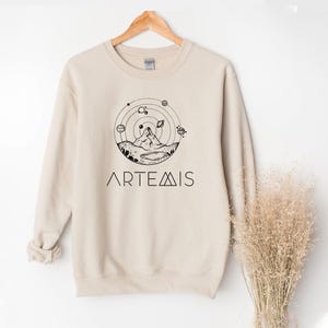 May include: A white ceramic mug with a black handle and the text "But First, Coffee" in a bold, black font. The mug is filled with a dark liquid, likely coffee, and sits on a light-colored surface.