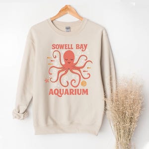 May include: Beige sweatshirt featuring a coral octopus design and the words "Sowell Bay Aquarium." The graphic includes fish, bubbles, and a seashell. The sweatshirt is hanging on a wooden hanger.