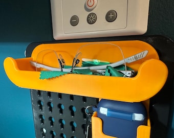 May include: Orange plastic wall-mounted organizer with a glasses holder and a small blue case. The organizer is attached to a black pegboard.
