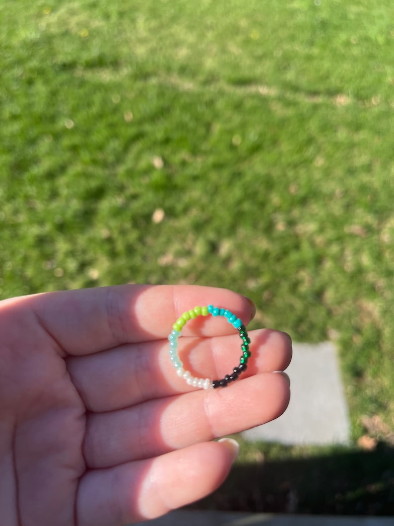 May include: A beaded ring with a gradient of green, blue, white, and black beads.