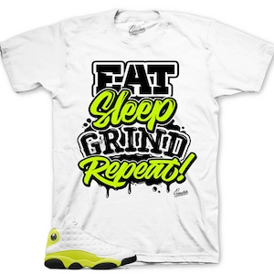 May include: White t-shirt with the text "EAT Sleep GRIND Repeat!" in black and neon green. A pair of white and neon yellow sneakers are in the bottom left corner. The shirt has a crew neck and short sleeves.