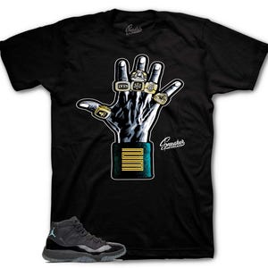 May include: Black t-shirt featuring a graphic of a hand wearing multiple championship rings. The hand is positioned above a teal and gold rectangle with the number 23. A pair of black and gray sneakers with teal accents are at the bottom.