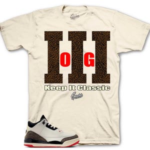 May include: Cream-colored t-shirt featuring a brown graphic with the letters "OG" in red, and the text "Keep It Classic." A pair of white, black, and red sneakers are in the foreground. The shirt has a crew neck and short sleeves.
