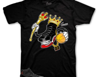 May include: Black and red Air Jordan 11 sneakers with a black t-shirt featuring a cartoon illustration of a sneaker wearing a crown and holding a basketball. The t-shirt has the text "Sneakers" on it.