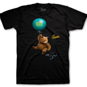 May include: Black t-shirt featuring a brown teddy bear holding a teal balloon with a gold design. Below the bear are black and teal sneakers. The shirt has the text "Sneaker Threads" in gold.