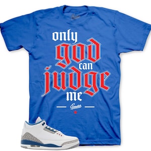 May include: A bright blue short-sleeve t-shirt featuring the text "only god can judge me" in a bold red gothic font with a white outline. Below the text is a small white logo and a red star. A white sneaker with grey elephant print accents.