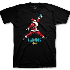 May include: Black t-shirt featuring a Santa Claus graphic in a jumping pose, holding a gift. The Santa is dressed in a red suit with a white beard and hat. The word "GAMMAS" is in teal below the Santa image.