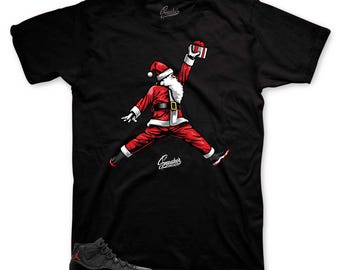 May include: Black and red basketball shoes with a Santa Claus graphic on a black t-shirt. The Santa Claus is wearing a red suit and is holding a gift box. The text "Sneakers" is printed on the t-shirt.
