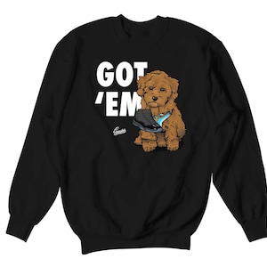 May include: Black sweatshirt with the text "GOT 'EM" in white. A cartoon dog is holding a black and blue sneaker. The sweatshirt has a crew neck and long sleeves.